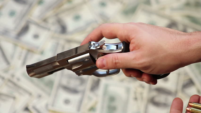 High angle of hands loading bullets into a handgun, over a background of hundred dollar bills.