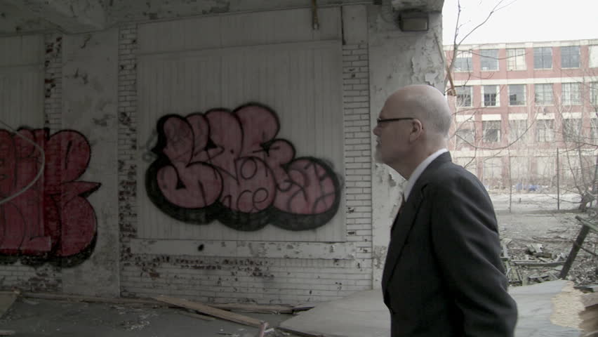Businessman walks past graffiti in broken down building, then out into the light. Symbolising decay, hope and opportunity, as well as light at the end of the tunnel. Hand held camera.