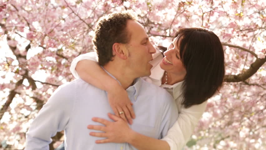 Mature man giving his wife a piggyback ride, both having fun and the loving couple kissing in the end with cherry trees in background