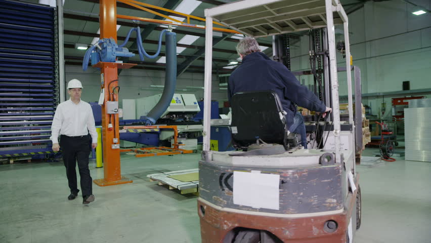 A team of workers in a warehouse or factory are going about their business and preparing goods for dispatch. One man in a forklift truck is lifting and moving stacks of wooden pallets. 