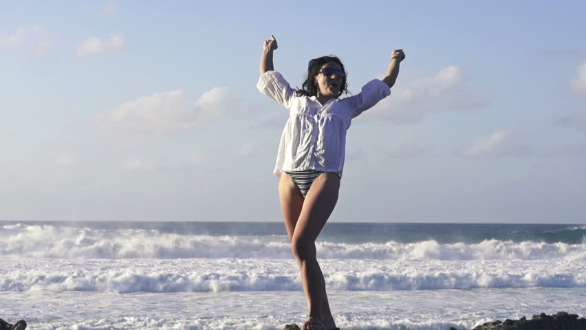 Happy, excited woman standing by the sea and raising arms in slow motion  
