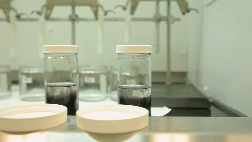 Two shots (one medium, one close-up) both using a slider (dolly) in a chemistry laboratory, moving past a row of glass jars that contain liquids.