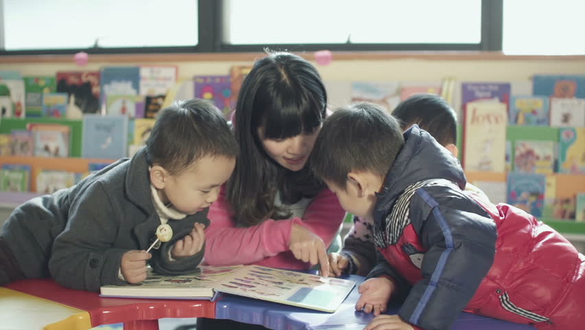 asian kindergarten teacher and her students sit a small table together together reading a book  in a classroom in china. 