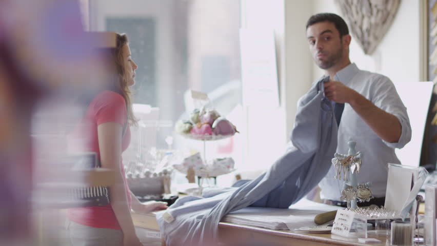 An attractive male store assistant or business owner chats to his female customer as he serves her. In slow motion.