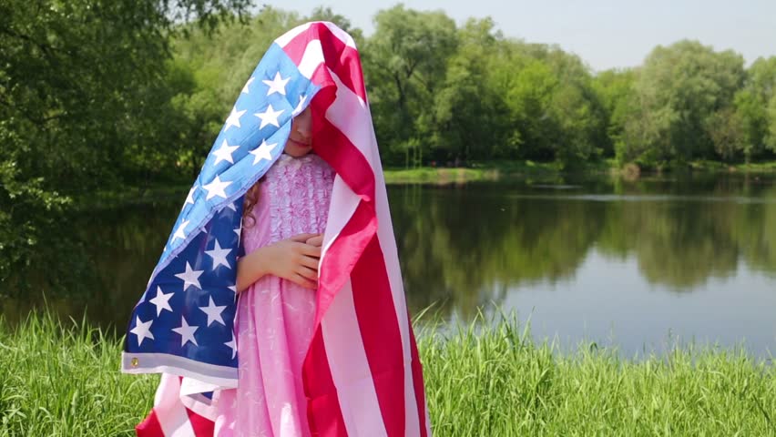 Little girl stands wrapped by US flag and then uncovers her face  with american flag near pond at sunny summer day in park