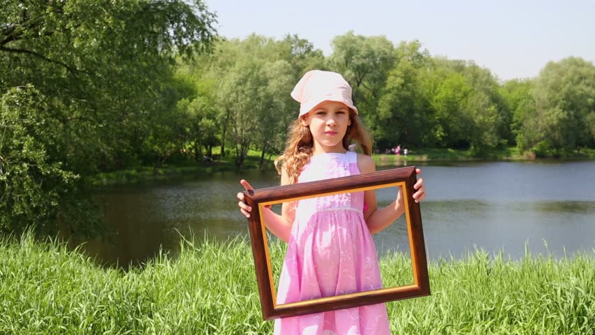 Little girl holds picture frame and shifts it sideward several times, near pond at summer park