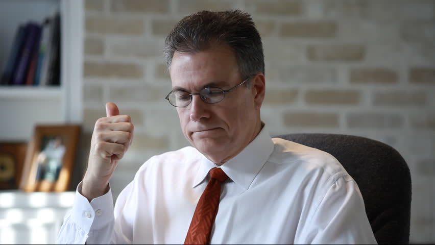 A man reading in his study looks up, removes his glasses, and smiles at someone off camera.