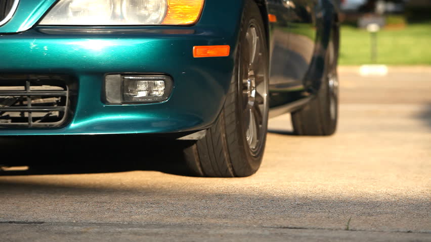 A sports car with fancy wheels, pulls up and a man in cowboy boots steps out onto the pavement.