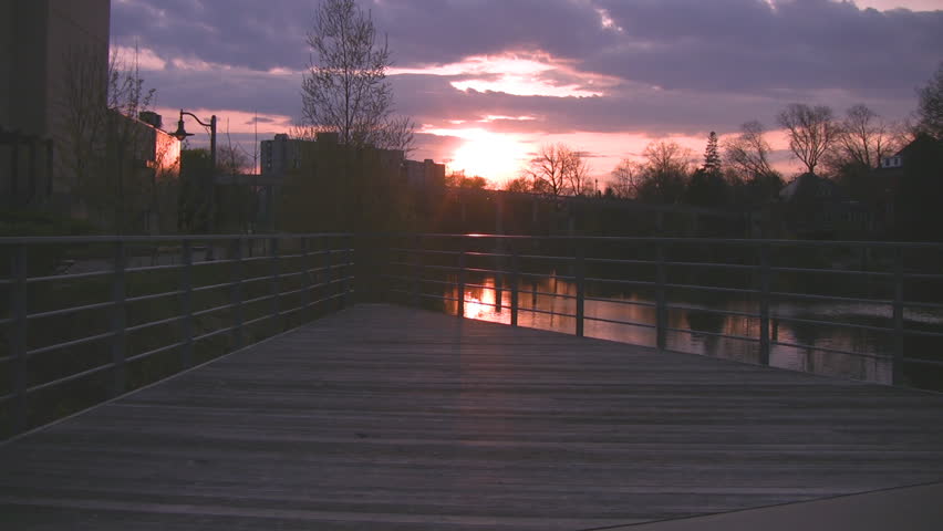 A view over a boardwalk with a setting sun in the distance.  The shimmering water of the Speed River in Guelph, Ontario, Canada, is also visible.