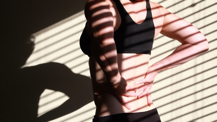 Light through window blinds frame a woman in workout attire who is experiencing back pain