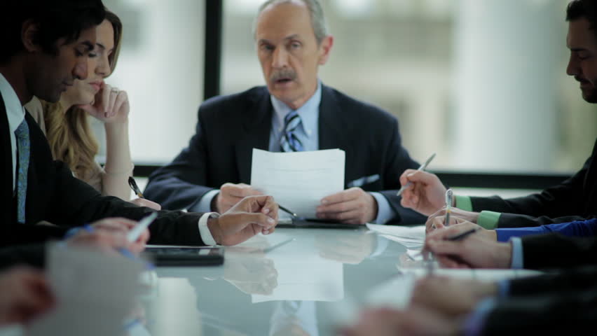 An older businessman leads a meeting at a large table and reads from his paper