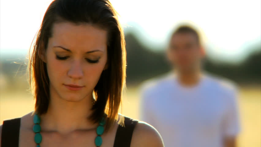 A young woman stands in a field and looks into the camera as her boyfriend stands behind her out of focus