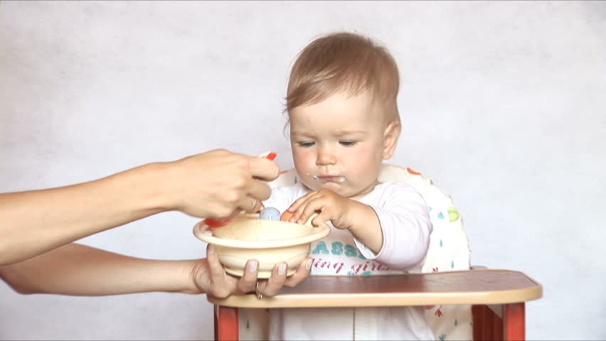 Baby sits at a children's little table. Mum feeds the child with porridge. Isolated on the white.