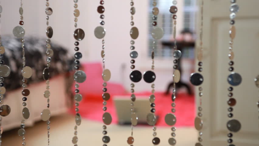 Two young teenage girls walk through decorative strands of beads and lay down on a pink rug and start looking at a laptop