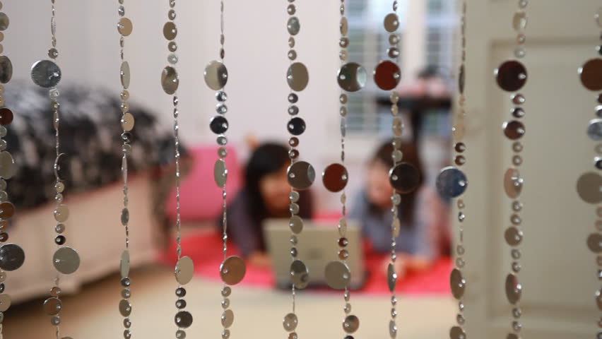 The camera rack focuses from decorative strands of beads hanging in a doorway to two young teenage girls laying on a pink rug working on a laptop.