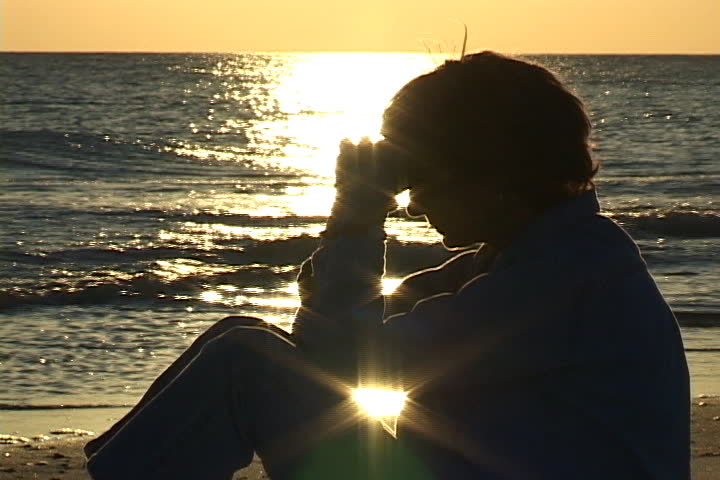 Mature woman sits on the beach praying with hands folded against head. Sunlight sparkles on the water in the background. Shot on miniDV from a tripod.