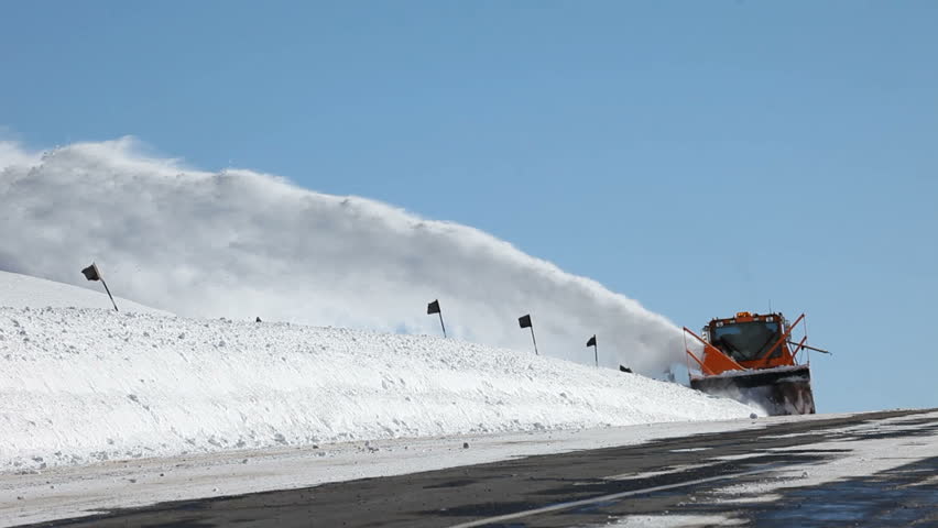 Snow blower removing snow from mountain road. On top of mountain road. Large vehicle with spinning blades throws ice and snow off the road. Deep and dangerous snow.