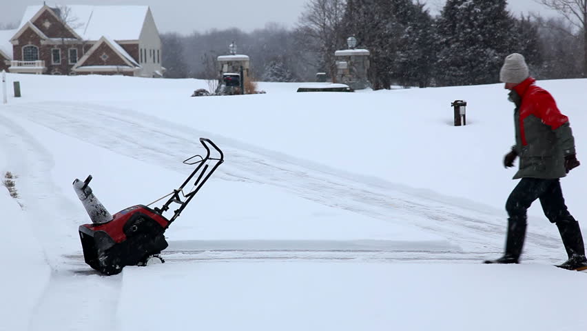 Senior man in red coat using a snow blower during a blizzard on home drive