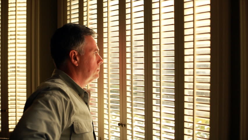 Afternoon sunlight pours in the shuttered windows on to a man who is gazing solemnly through them.