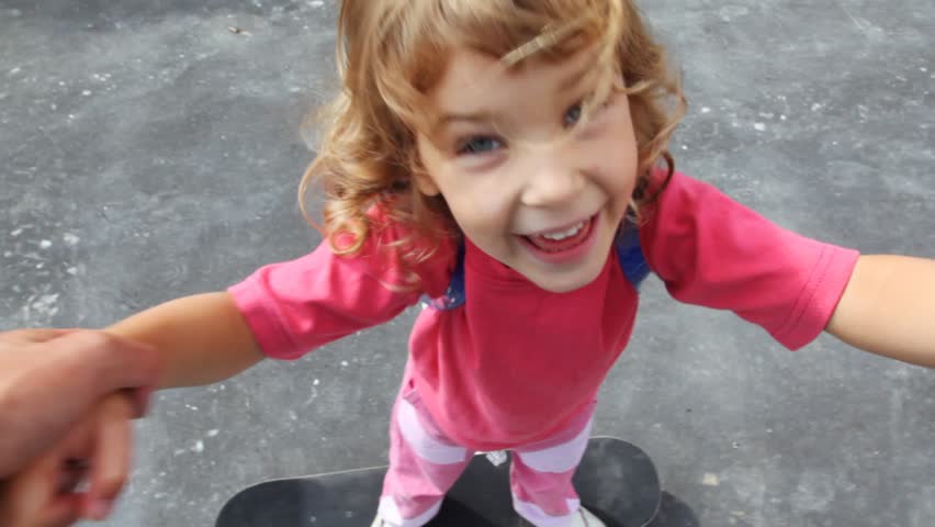 little girl stands on skateboard and it roll on it holding for hands on asphalt 