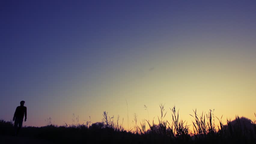 silhouette of traveler with backpack walks downhill against sunset sky to camera 