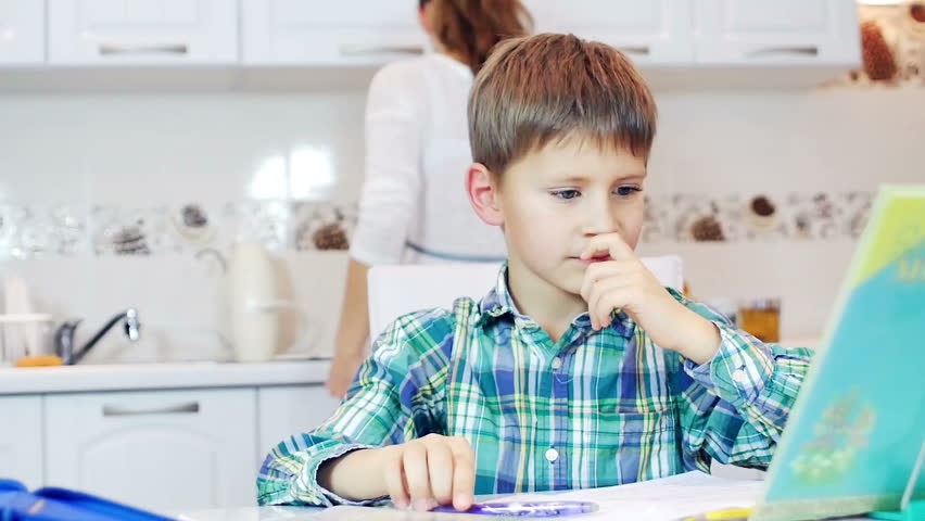 Little Boy Doing Homework In The Kitchen While His M