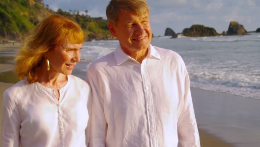 Older couple hold hands and walk on the beach together. 