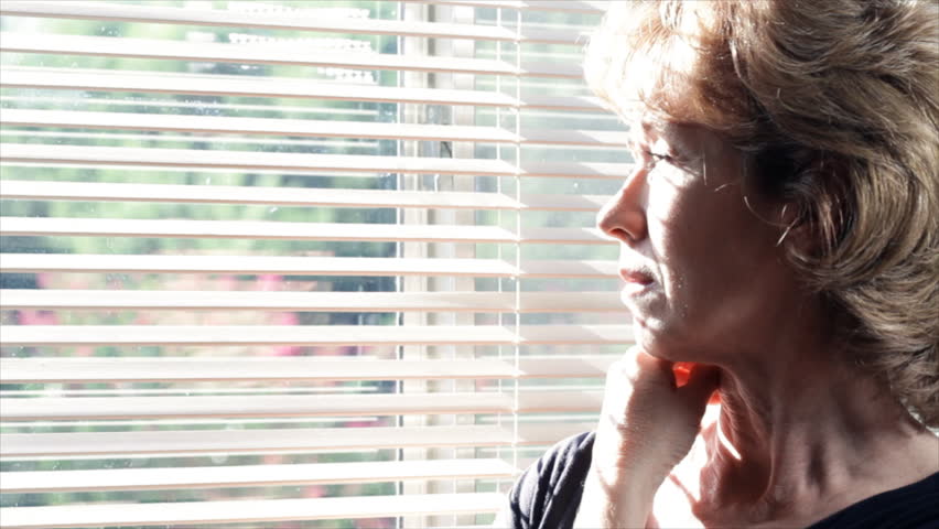 An older woman gazes out a sunlit window then turns and looks past the camera.