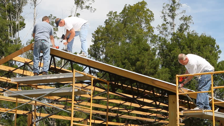 Construction crew uses powered screw guns to attach a sheet metal roof to the rafters of a barn.