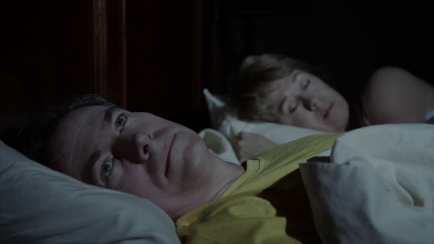 A sleepless man lies wide awake in bed while his wife is sleeping soundly next to him.