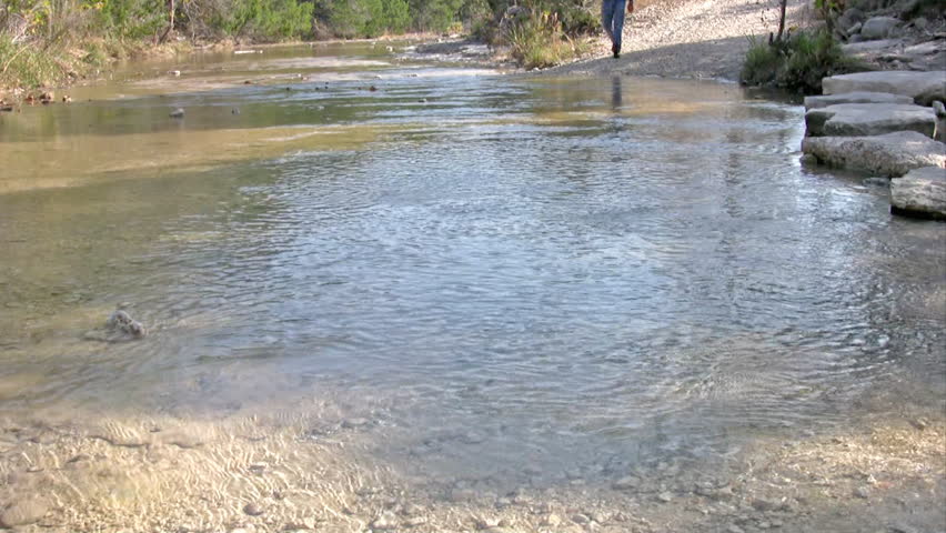 Video of a man walking across a shallow stream towards view. Legs and boots only.
