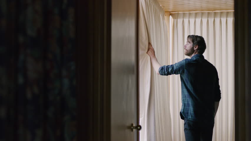 Young bearded man opening window curtains in a dark room. Shot with a RED camera. 4k footage.