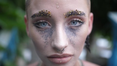 Close Up Of Woman With Sparkling Glitter Makeup Facing The Camera Portrait Of Lady With Shaved Head And Nose Ring Standing Outside Gives Sly Smile While Looking Into Camera Light Blue Eyes