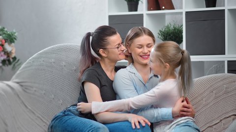 Same sex female family smiling hugging daughter sitting on couch looking at  camera