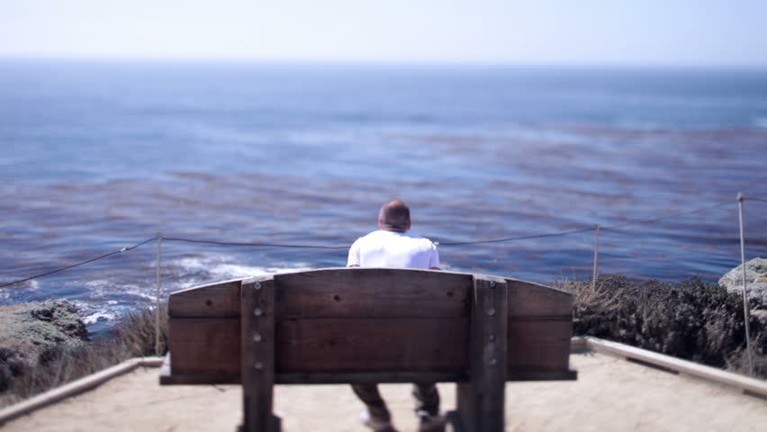 A Man Takes A Seat On A Wooden Bench At Big Sur California Looks
