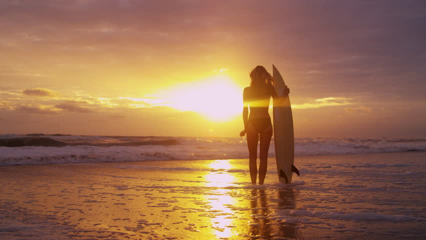 Young Female Surfer Standing On Beach Sand Under Sunset Sky Holding ...