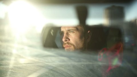 Man is driving a car. Reflection face in rearview mirror of vehicle. Time of sunset in a city